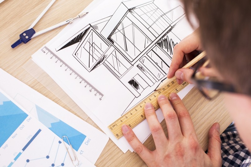 Top view of young architect using ruler and pencil to draw blueprint on wooden desktop with business report and a pair of compasses